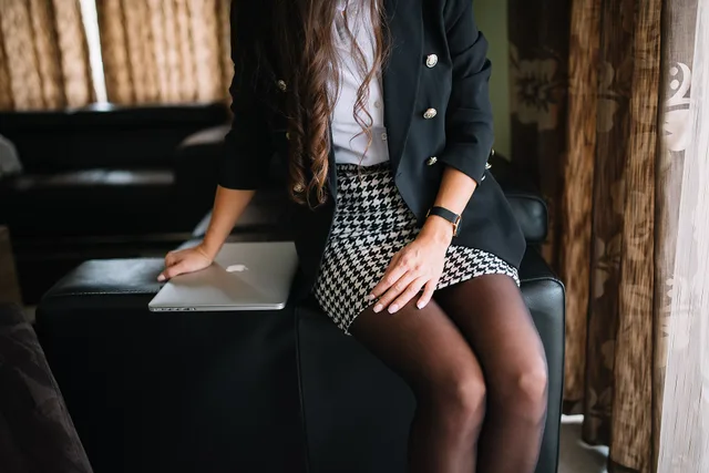 A young woman sits on a black couch in a modern living room, wearing a blazer and a fashionable skirt while working on a laptop. Sunlight streams through the curtains. Business professional attire for women: the three-piece test that works
